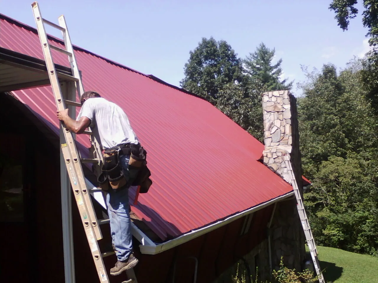 Roofer on a ladder beside a red metal roof and stone chimney — Soffit & Fascia Repair by Roofing Brothers in Kendalia